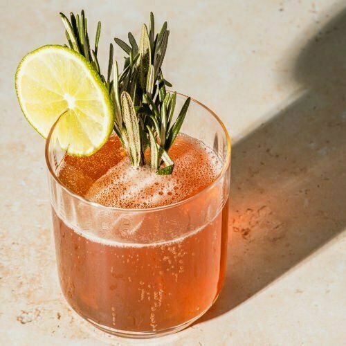 Pink cocktail in small whiskey tumbler and garnished with a slice of lemon and a sprig of rosemary