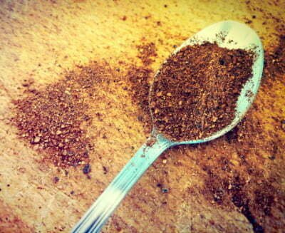 An antique metal teaspoon lies heaped with ground nutmeg.