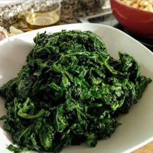 A large bowl of cooked spinach sits on a kitchen countertop with some olive oil, garlic, and chickpeas sitting in the background