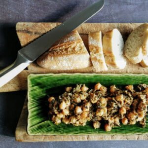 A long green plate of spinach with chickpeas sits on a rustic chopping board with a few slices cut from a white laof of bread