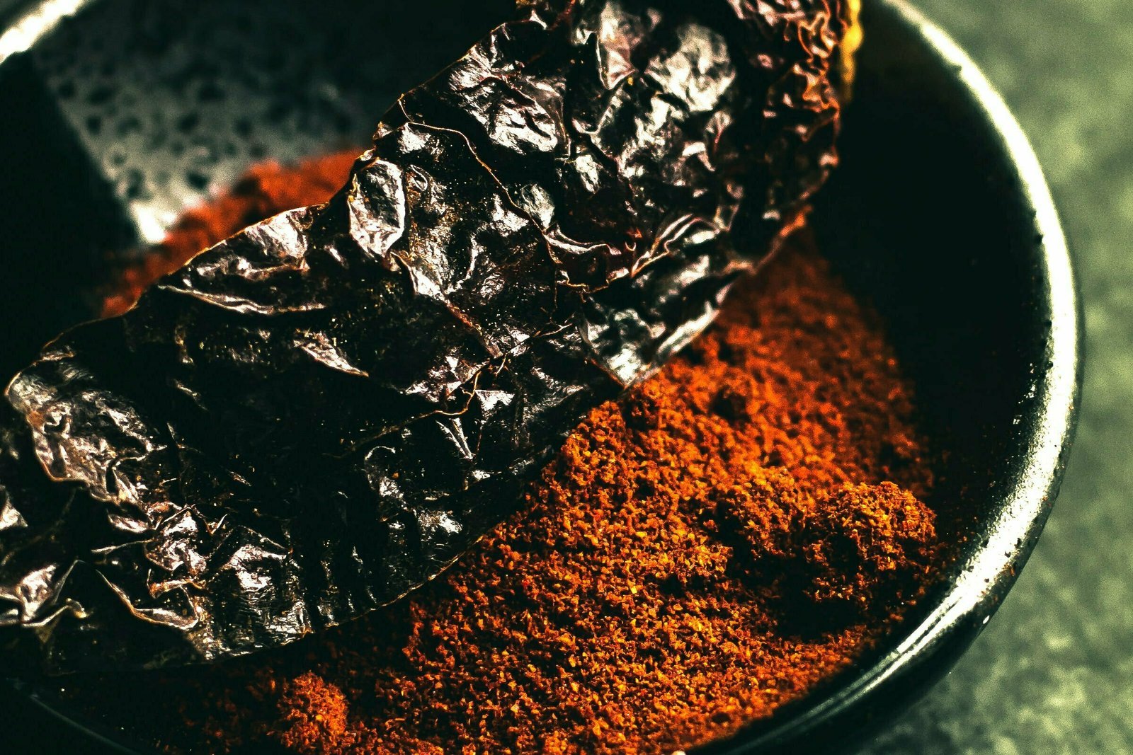 A rustic bowl of sweet paprika sits on a kitchen table. A large dried pepper sits on top of the paprika bowl. 