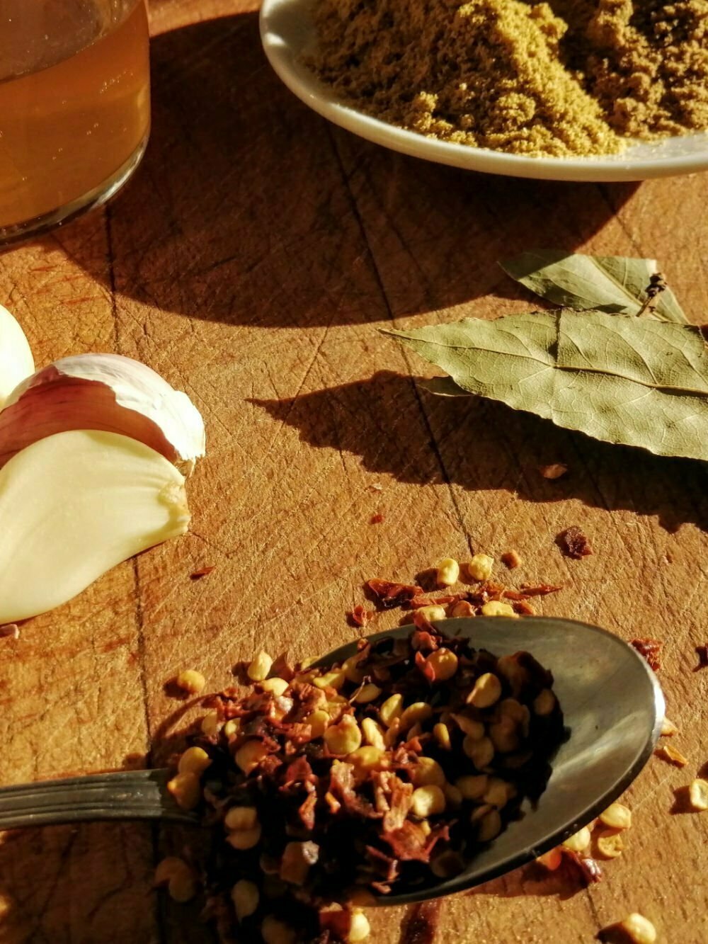 A rustic choppig board sits loaded with ingredients for making spinach with chickpeas, including dried chili flakes, bay leaves, garlic, and grund corriander