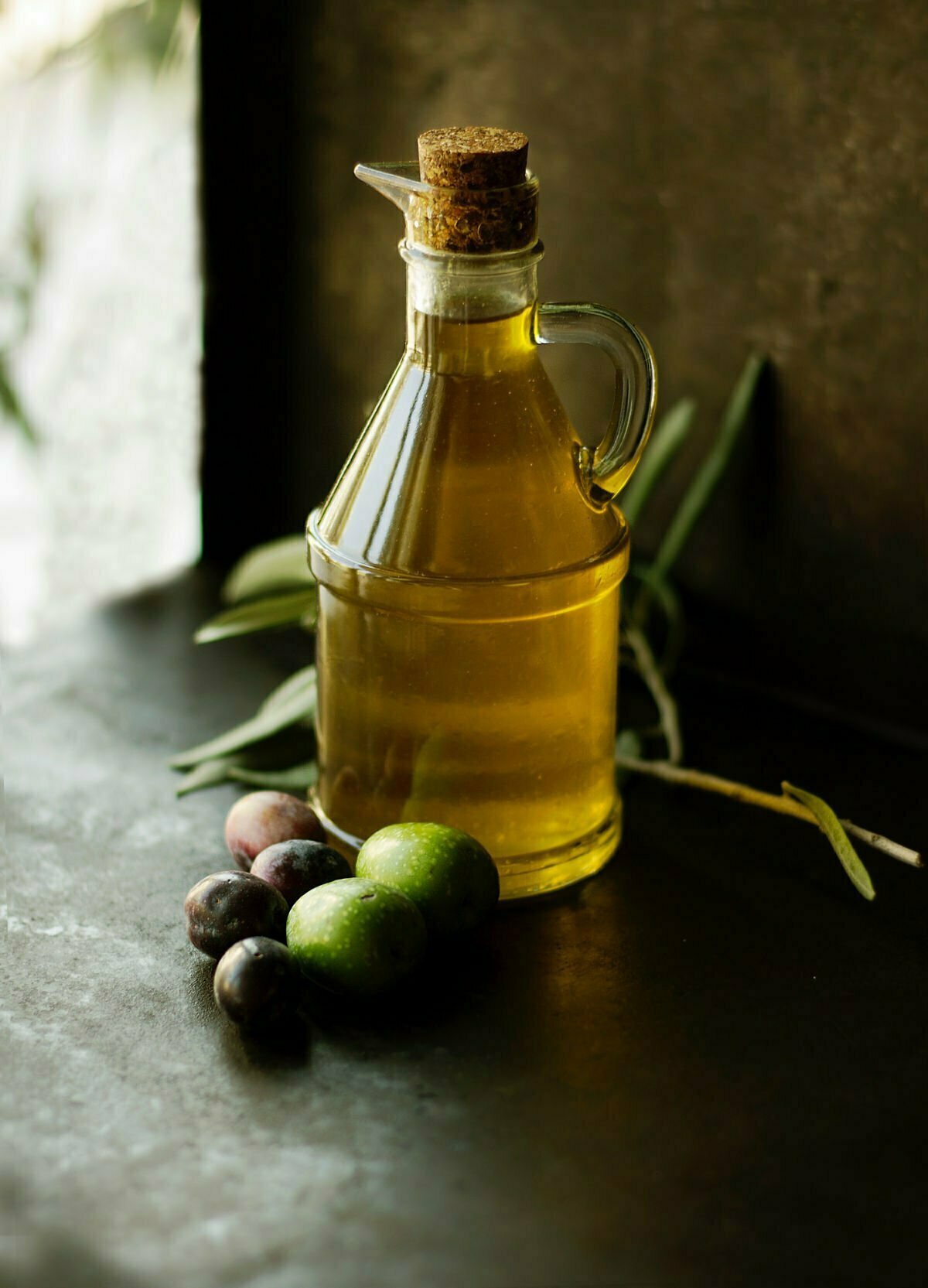 a large jug of olive oil sits on a rustic benchtop with a few fresh olive branches beside the jug
