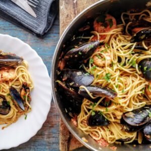 A large pot is filled with Shrimp and Mussels pasta and sits beside a small bowl topped with pasta, cooked mussels, shrimp, and cilantro