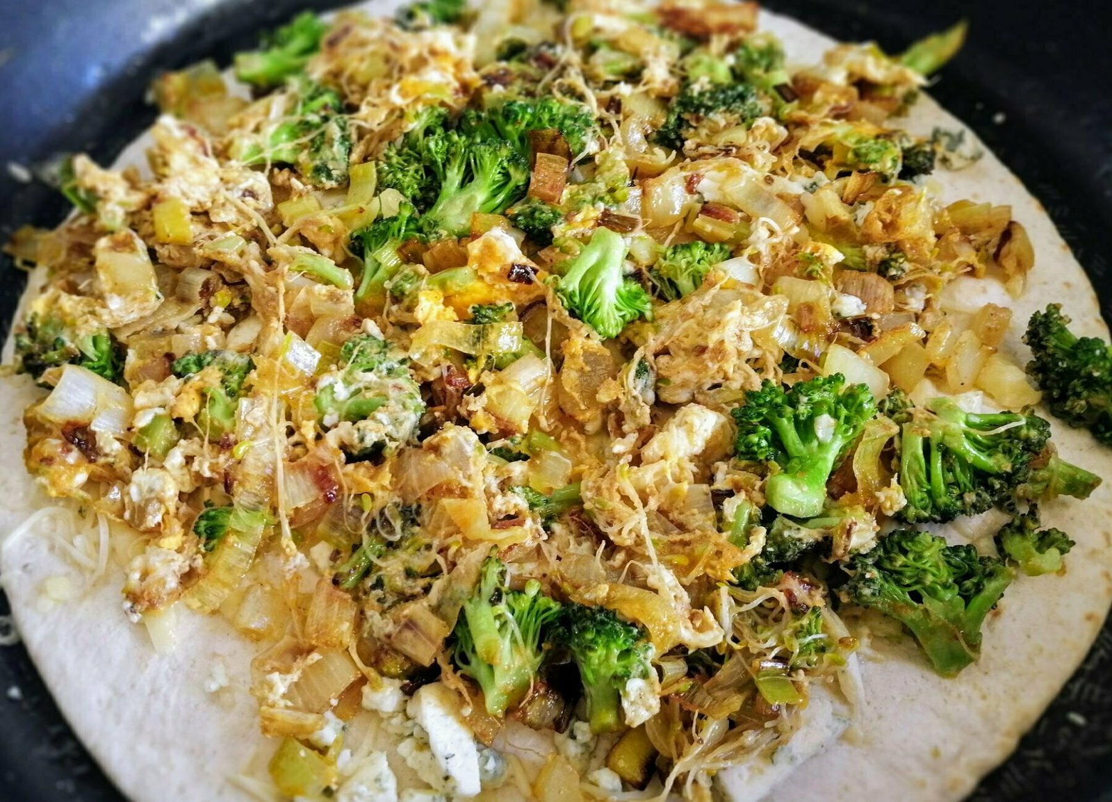 An opened quesadilla sits in a pan with leek brocolli and blue cheese sprinkled on top