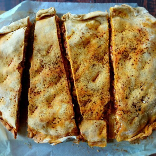 A large empanada gallega sits cut up into peices on some parchment paper
