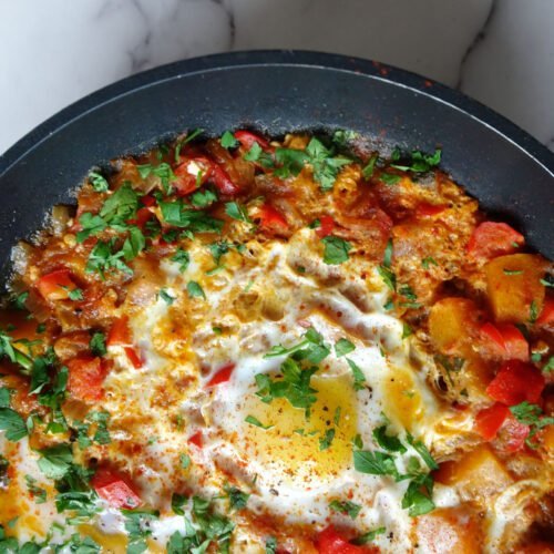 A frying pan of vegetable pisto with eggs sits on a white marble countertop