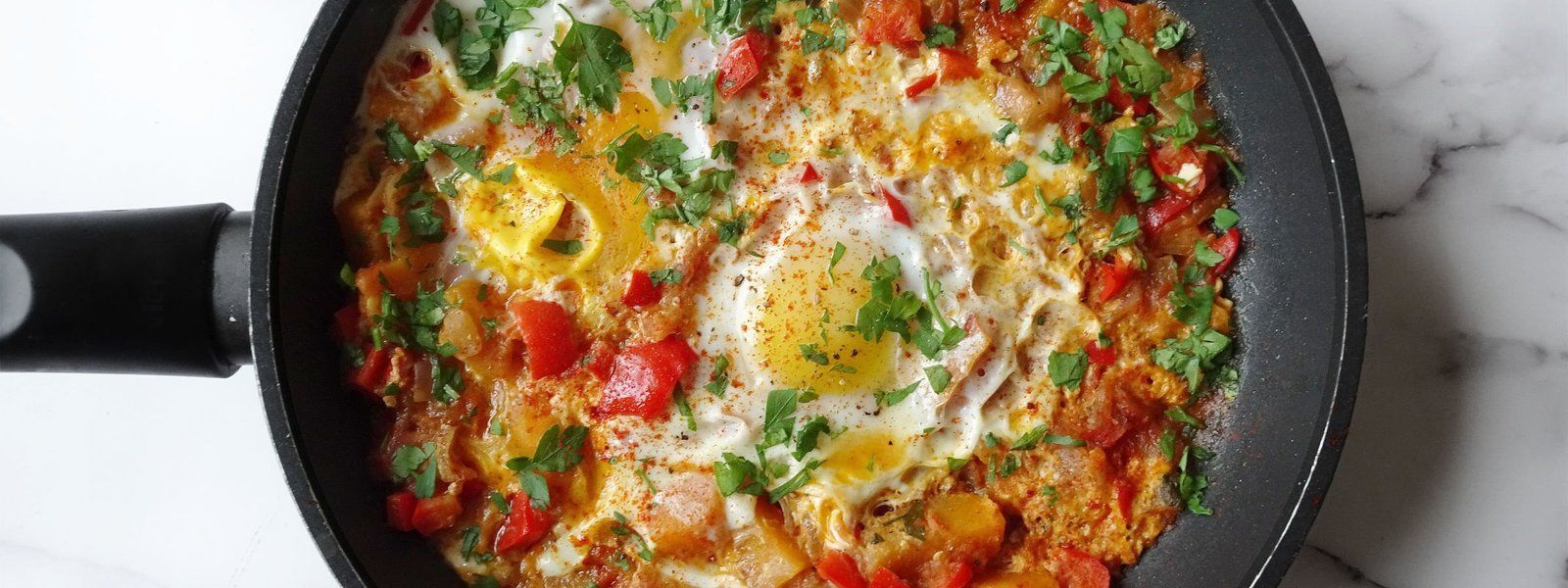 A frying pan of vegetable pisto with eggs sits on a marble counter