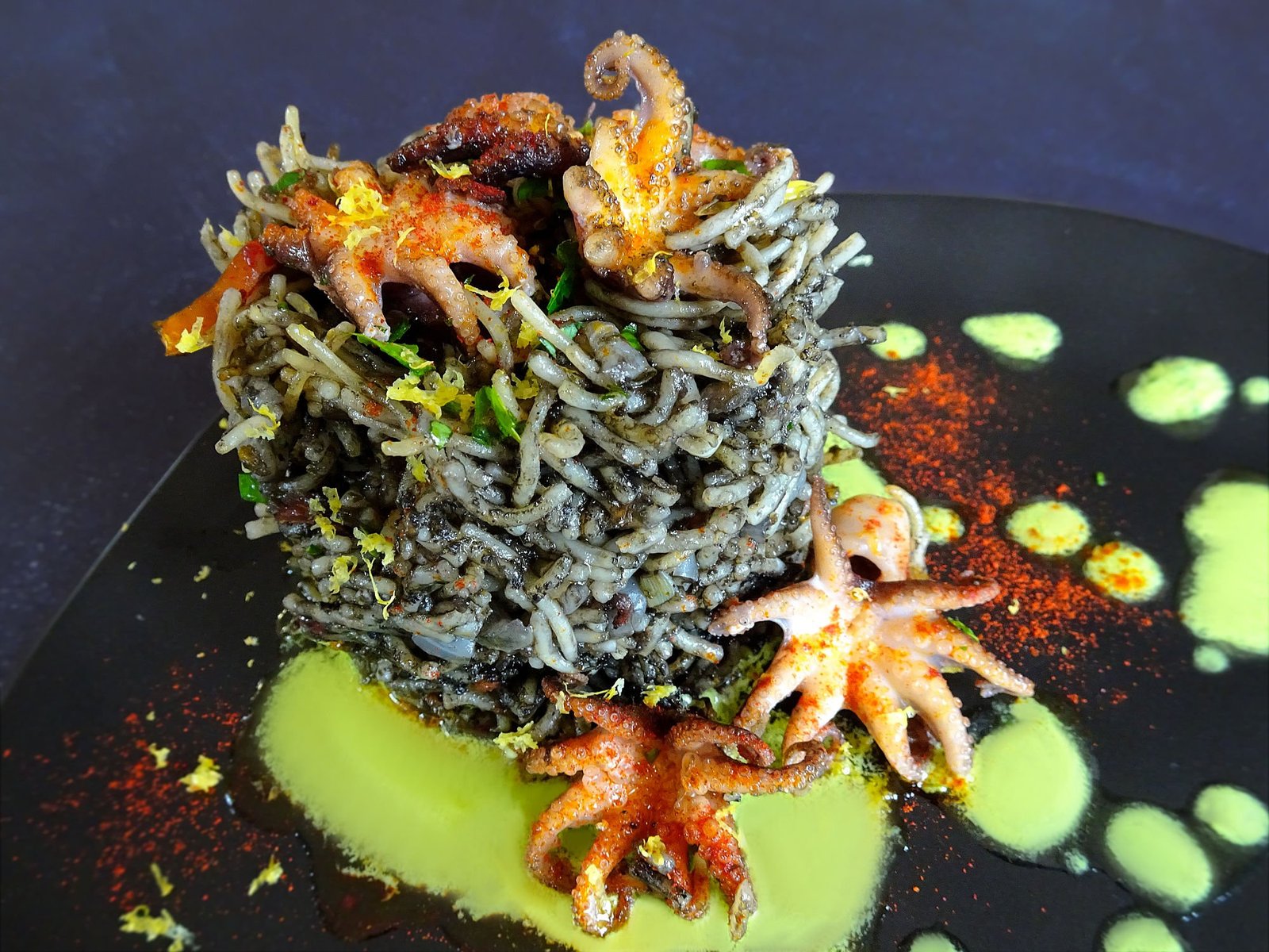 A tower of fideua pasta sits on a plate topped with green allioli and red baby squid