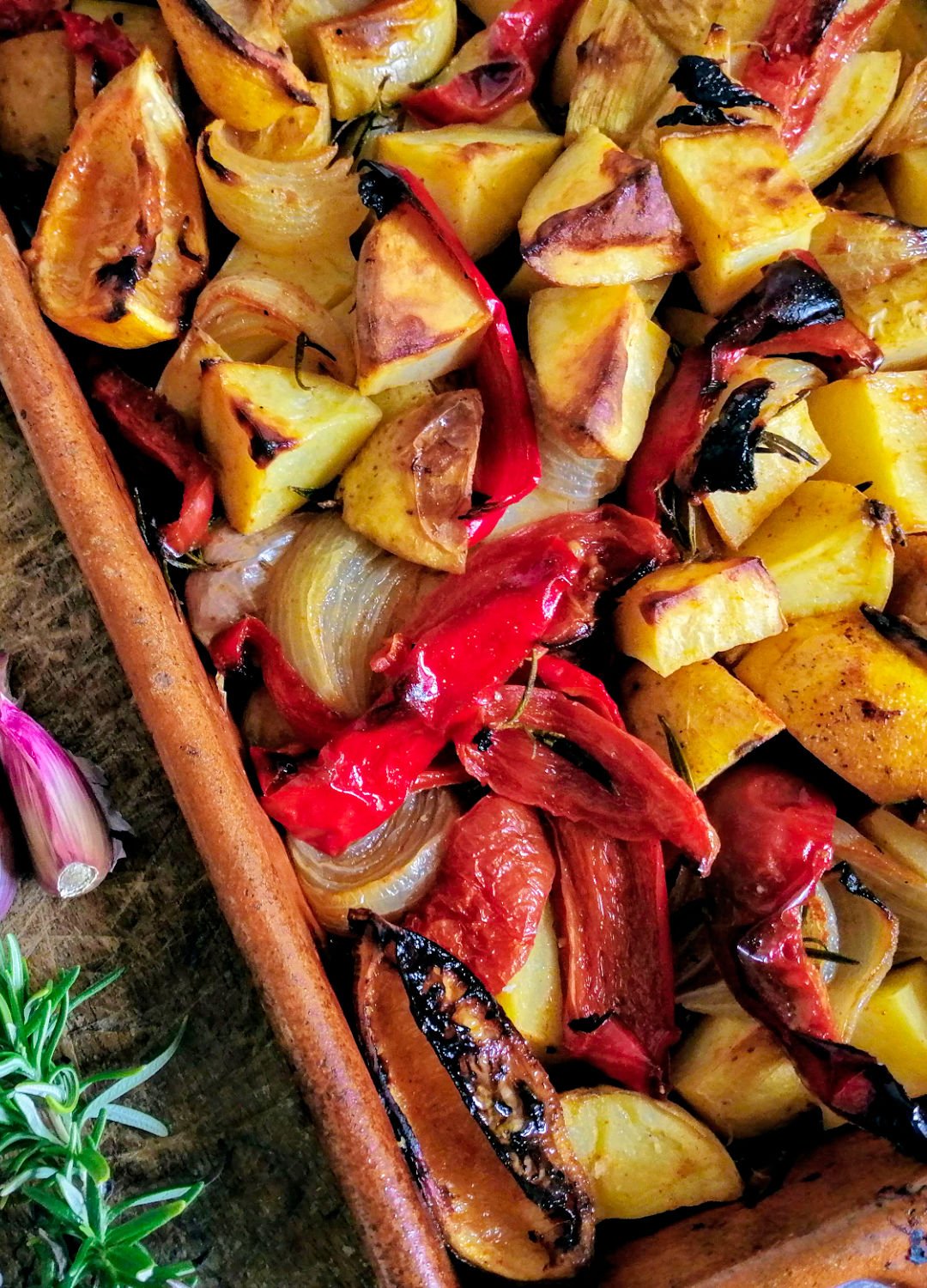A large earthenware dish sits filled with poor mans potatoes beside a few sprigs of rosemary and ssome garlic