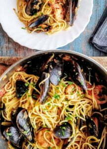 A large pot is filled with shrimp and mussels pasta and is being served onto a white plate in the background