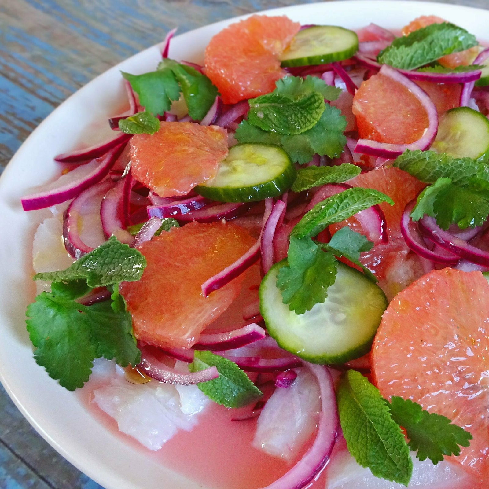 White fish ceviche sits on a white plate garnished with finely sliced red onion, pink grapefruit wedges, and some sprigs of mint and cilantro
