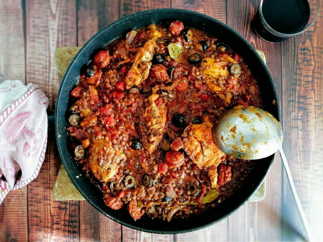 A large pan of chicken and chorizo stew sits beside a glass of red wine and a rustic napkin