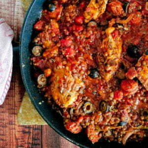 A large black pan sits on a rustic wooden board and is filled with chicken and chorizo stew