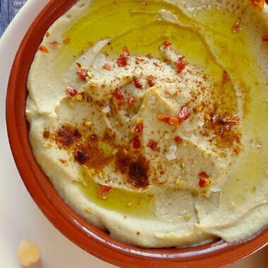 An earthenware bowl sits filled with Mediterranean hummus thats is topped with olive oil and some paprika