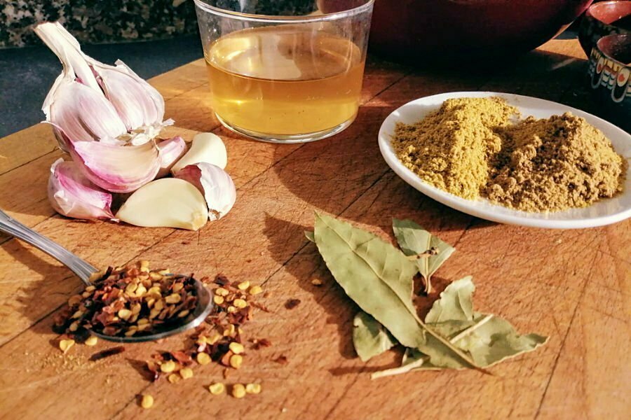 a chopping board sits with some garlic chili flakes bay leaves and a plate of spices near some olive oil in a glass