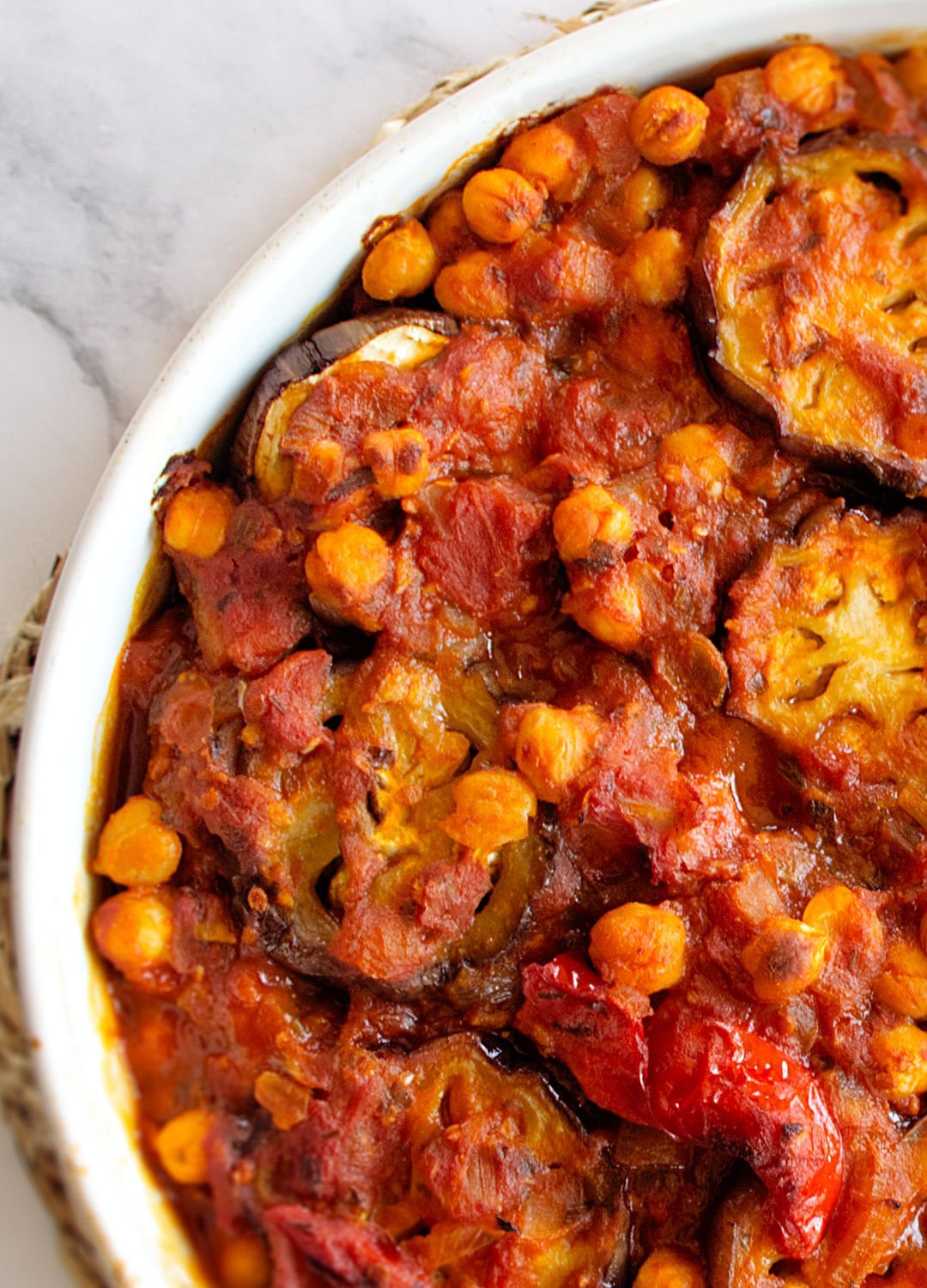 A large white casserole dish sits on a white marble counter with a rich red Mediterranean Eggplant casserole with chickpeas and tomato