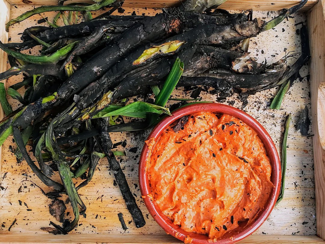 Barbercued calcots sit on a wooden servig tray with a small bowl of romesco sauce