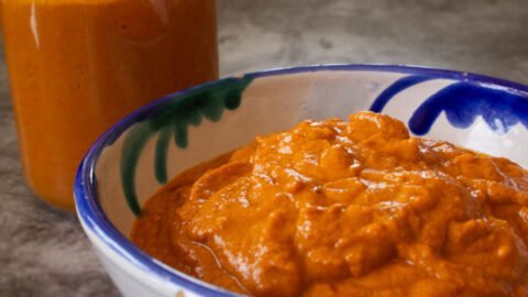 A small bowl of romesco sauce sits with a jar full of romesco sauce in the background