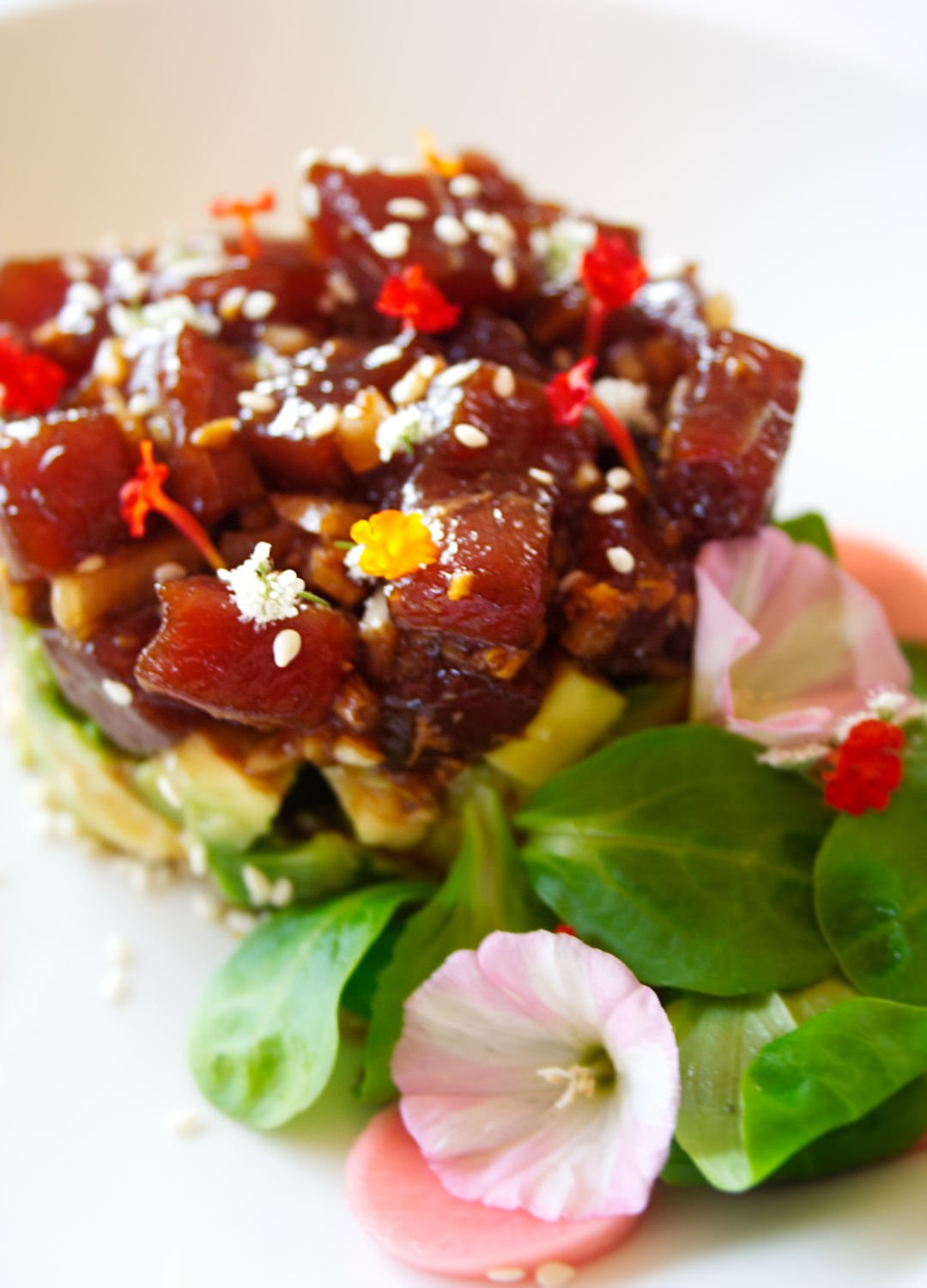 A stack of tuna tartare is built on top of some avocado and garnished with some green salad leaves and edible flowers