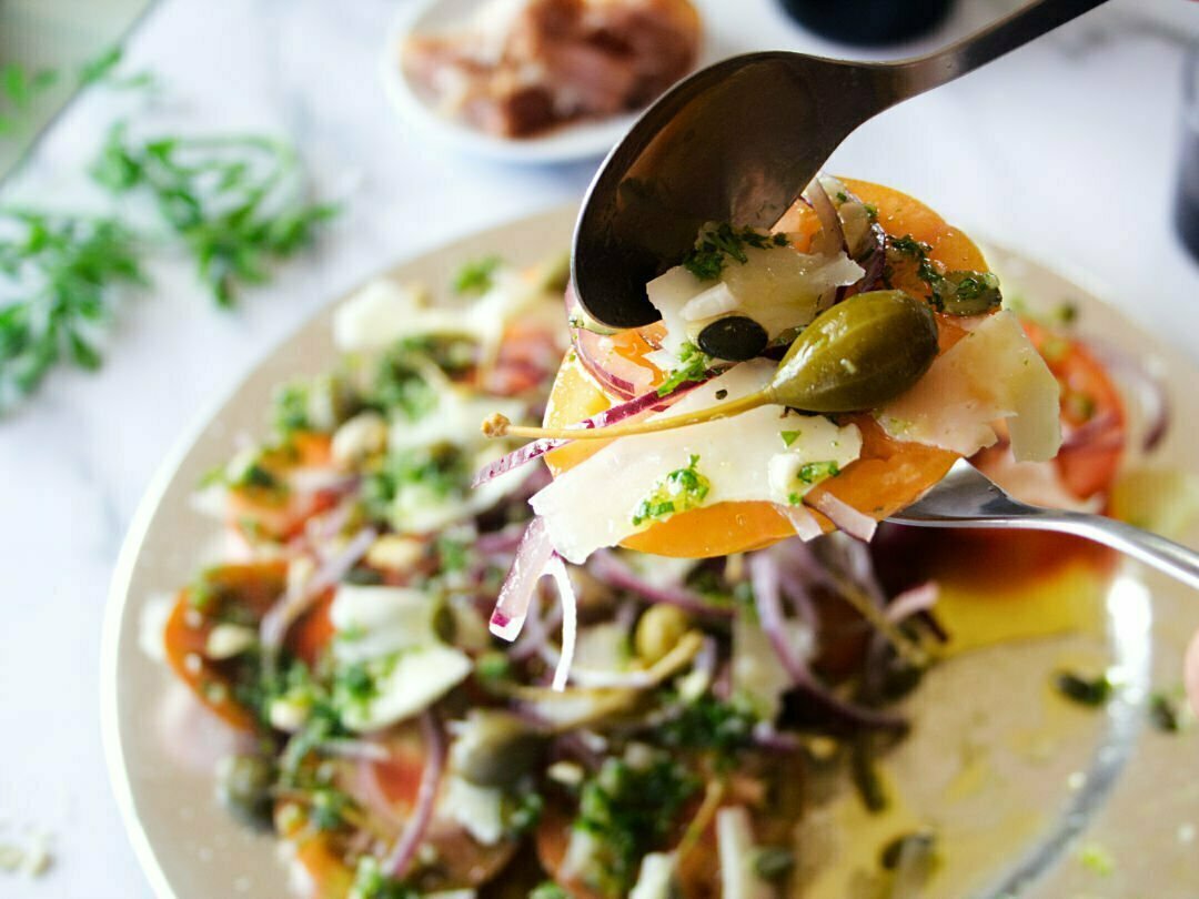 A spoonful of Mediterranean Tomato Salad is held above a large salad platter