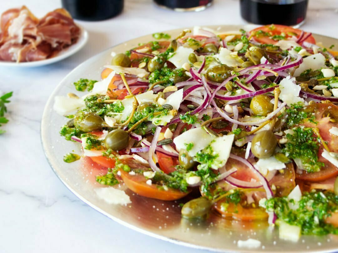 A large plate of Mediterranean Tomato Salad sits with lots of pesto dressing and shaved pieces of Manchego cheese