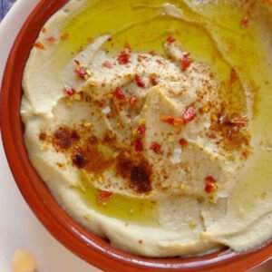 A small earthenware dish sits filled with Mediterranean Hummus and garnished with some paprika and olive oil