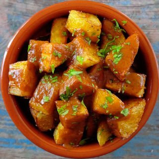 A small earthenware dish sit filled with patatas bravas and garnished with some freshly chopped parsley