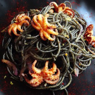 Squid ink pasta is served in a balck plate and garnished with some bright red baby octopus