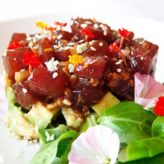 a small stack of tuna tartare sits on a white plate beside some green salad and flower garnishing