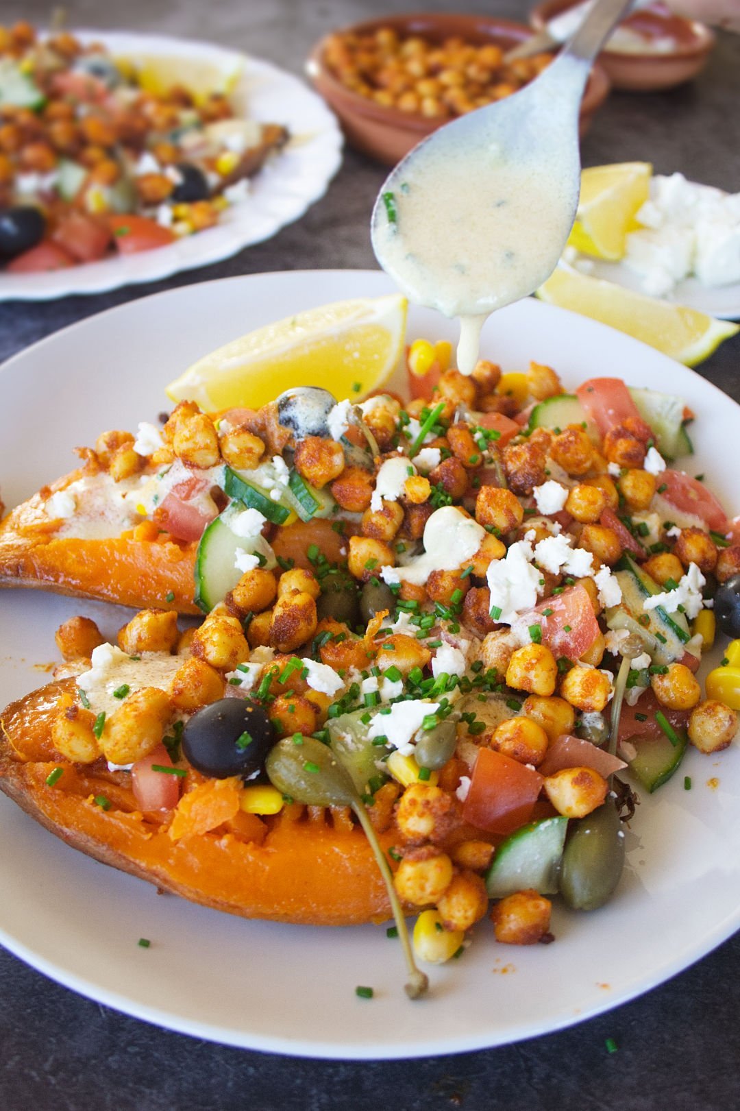 a large baked sweet potato sits on a white plate with plenty of Mediterranean inspired toppings