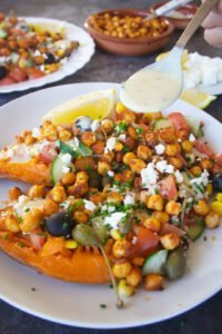 a large baked sweet potato sits on a white plate with plenty of Mediterranean inspired toppings