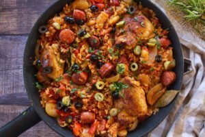 A large pan of Spanish chicken and rice sits on a counter beside some fresh rosemary