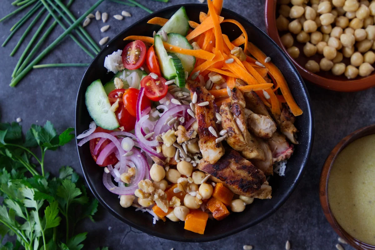 A grilled chicken rice bowl sits on a table with plenty of herbs and sald toppings