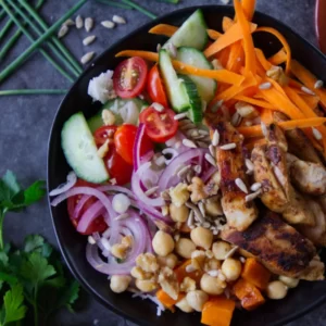 A grilled chicken rice bowl sits on a table with plenty of herbs and sald toppings