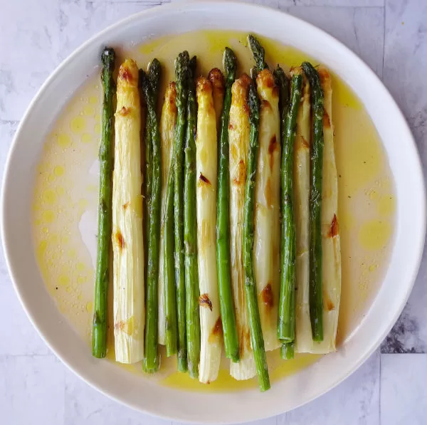 a large white plate sits on a marble counter stacked with roasted stems of white and green asparagus A pool of oil and white wine vinegar dissolves around the stack of roasted asparagus