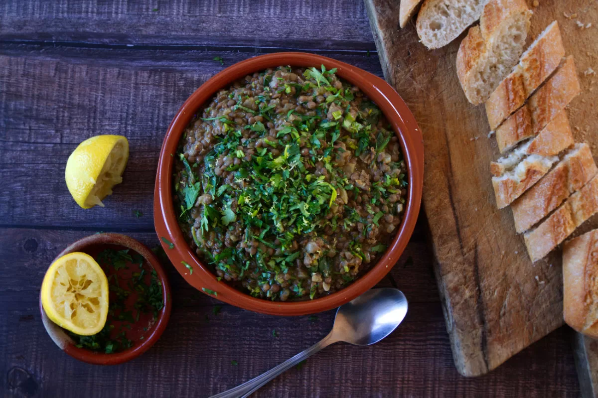 A bowl of lentil stew sits beside a bread bboard with some sliced bread.