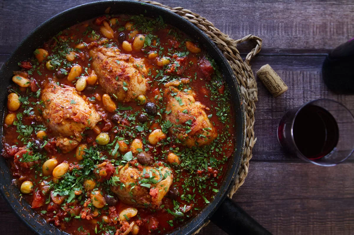 A pan of Spanish chicken thighs with chorizo and wine sits beside a small glass of red wine