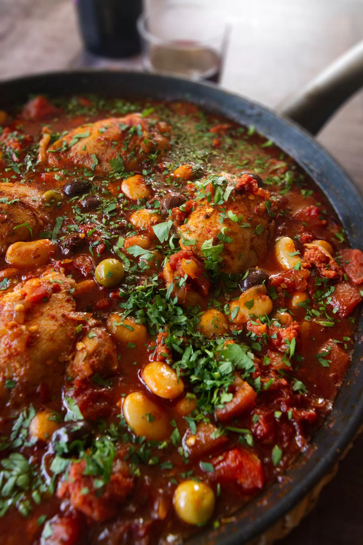 A pan of Spanish chicken thighs with chorizo and wine sits beside a small glass of red wine.