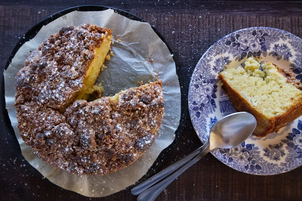 An pple cake is dusted with icing sugar and a slice is served. 