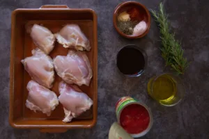 Uncooked chicken thighs sit in a dish beside tinned tmoatoes, herbs, spices, and red wine.