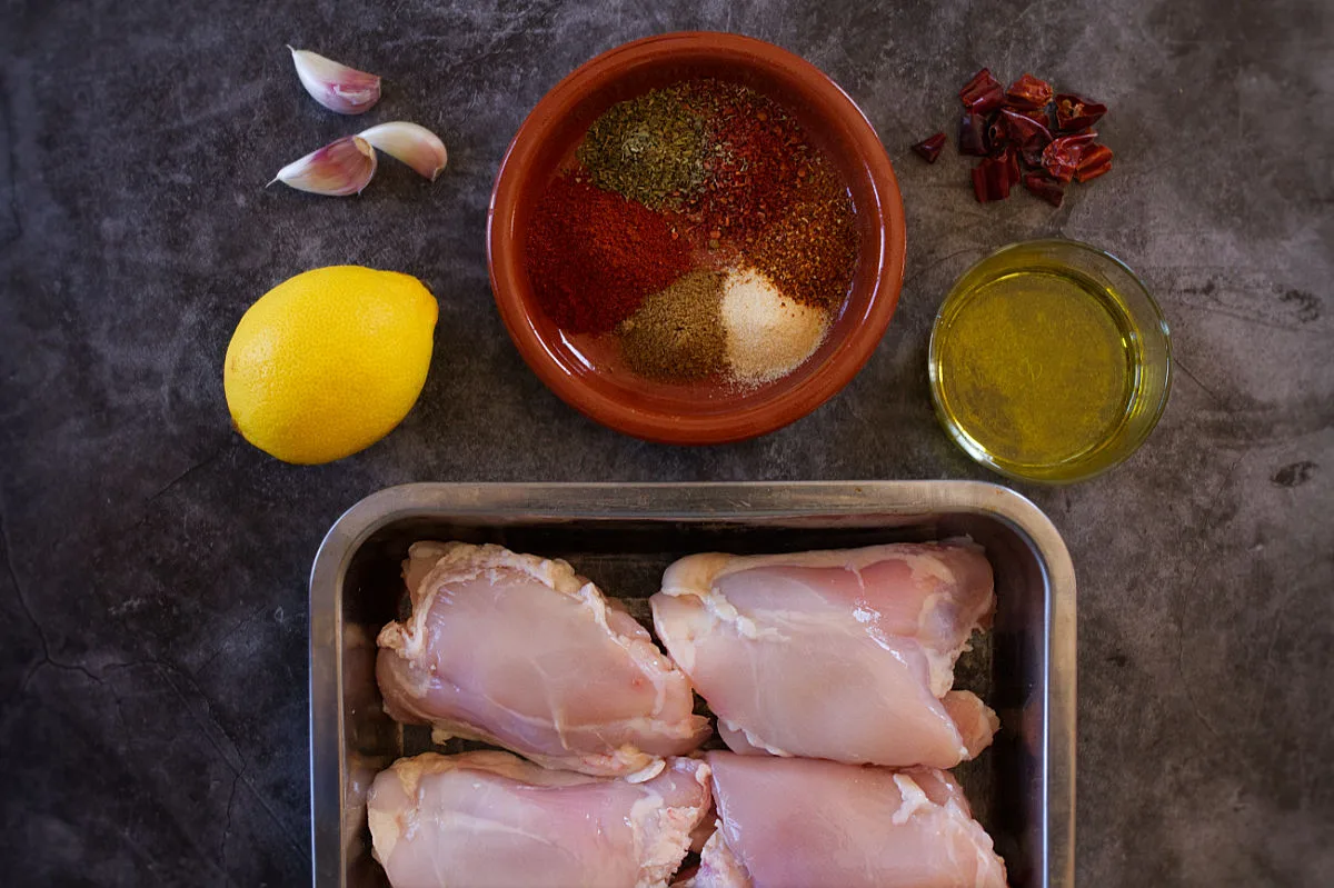 Some uncooked chicken thighs sit in a baking tray beside spices, a lemon, and some olive oil. 