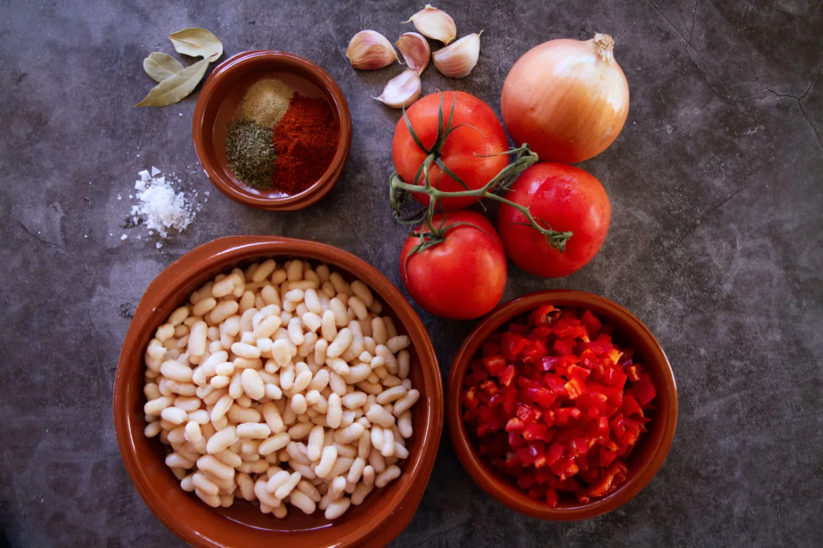 A large bowl of cannellini beas sits beside some tomatoes, onion, and spices used to make a Spanish-style white bean stew. 