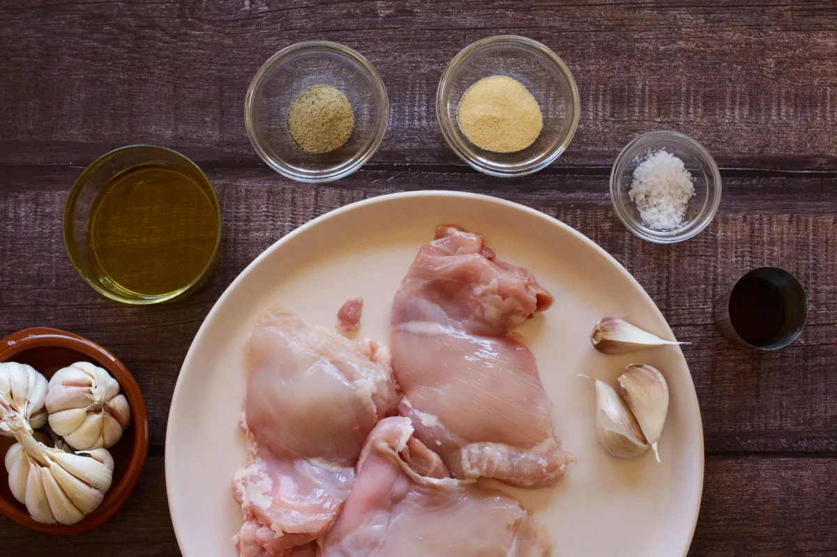 Some uncooked boneless chicken thighs sit on a plate beside some spices and garlic. 