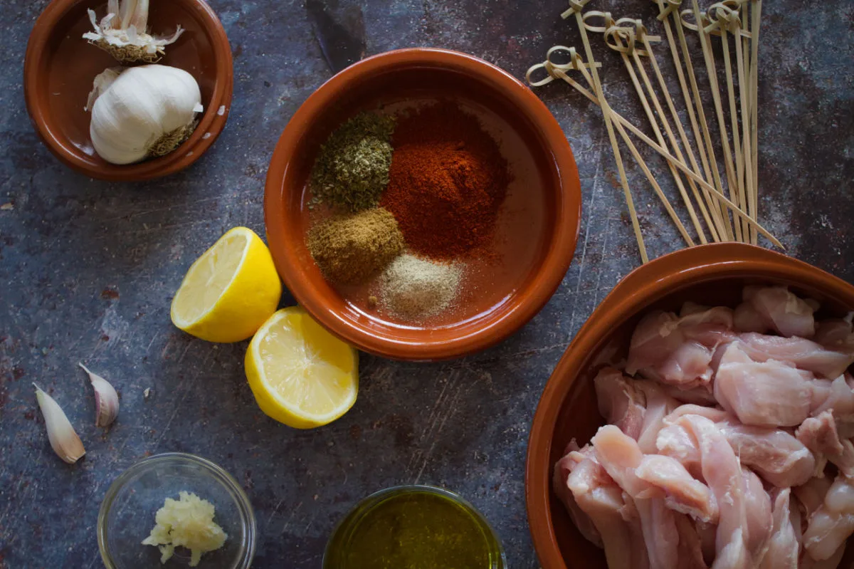 spices, paprika, lemon, garlic, and chicken pieces sit beside some skewers. 