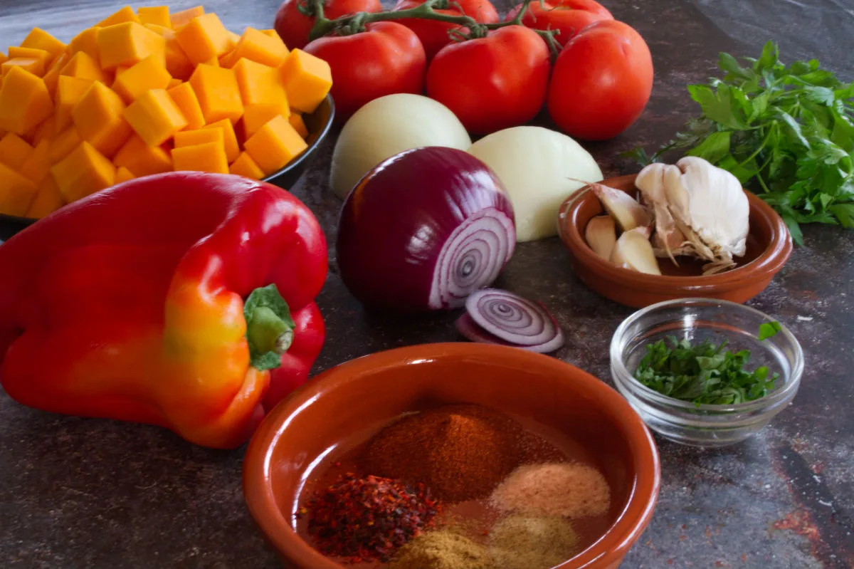 Some red bell pepper, pumpkin, onion, and spices sit on a counter. 