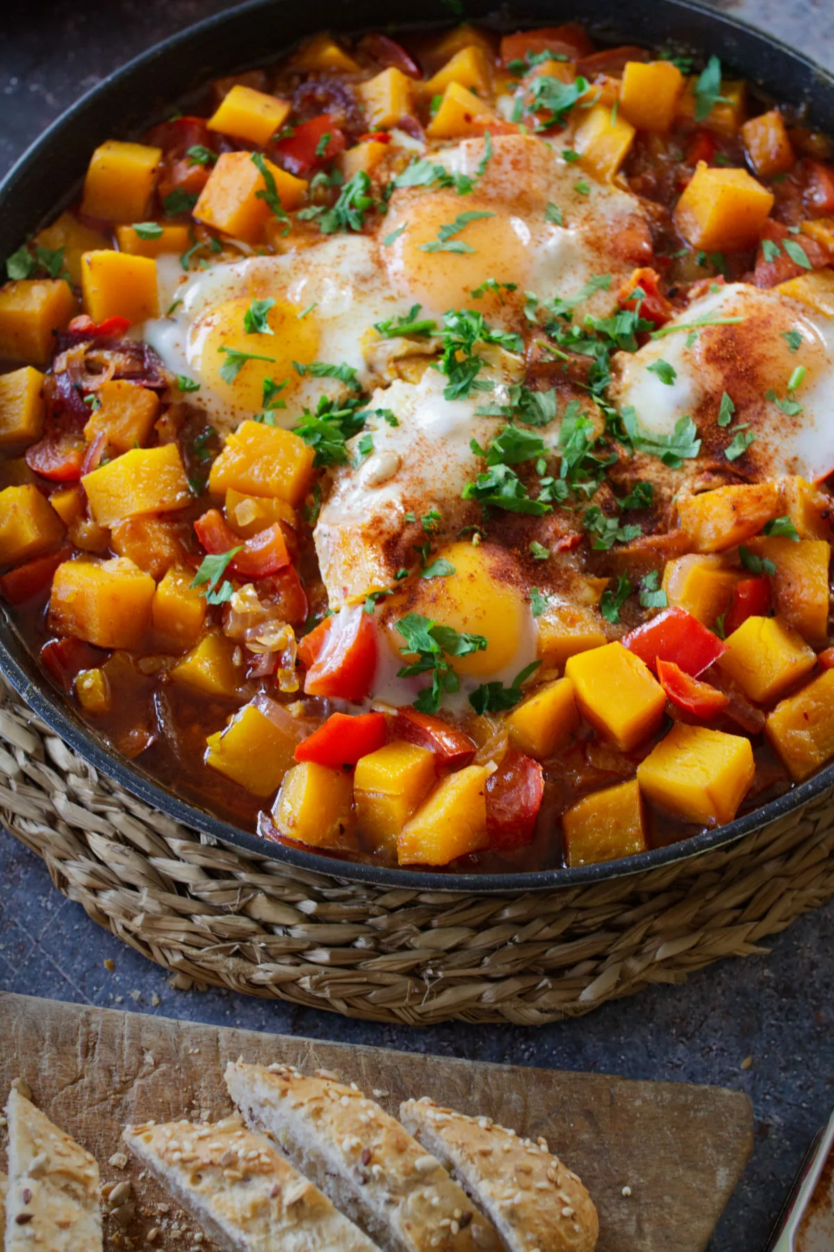 A large pan of pumpkin pisto sits beside some brown bread and fresh parsley.