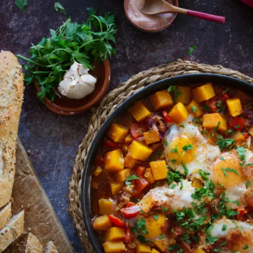 A large pan of pumpkin pisto sits beside some brown bread and fresh parsley.