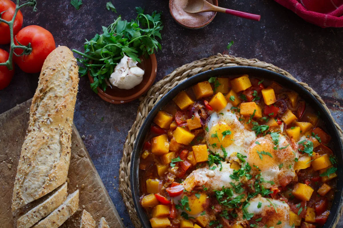 A large pan of pumpkin pisto sits beside some brown bread and fresh parsley.