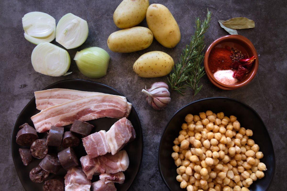 A few different cuts of pork and some sliced morcilla sausage sits beside some potatoes, onions, garlic, and spices. 